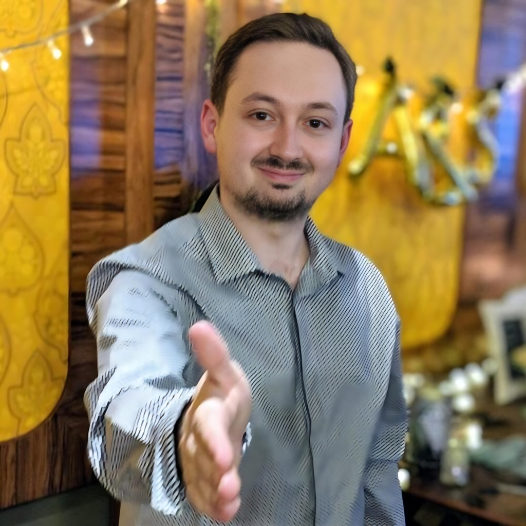 Man in a patterned shirt extending his hand for a handshake, smiling in a warmly lit indoor setting.